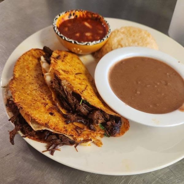 Two crispy birria tacos with a side of refried beans and a small bowl of consommé.
